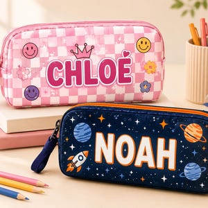 Peut inclure: Deux trousses rectangulaires avec fermetures éclair et dragonnes. La trousse du haut est rose avec un motif à damier blanc, des visages souriants, des fleurs et le nom CHLOE. Celle du bas est bleu foncé avec des planètes, des étoiles, une fusée et le nom NOAH.