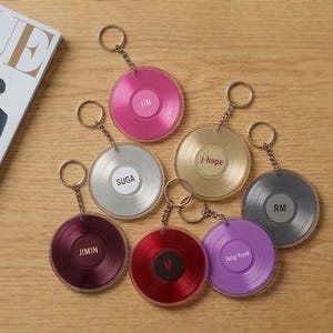 May include: A collection of colorful vinyl record-shaped keychains with gold-colored keyrings. Each keychain features a different color and a name printed on the center label. A magazine with the word "VOGUE" is visible in the background.