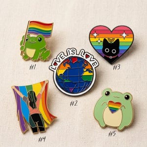 May include: Five enamel pins featuring pride themes: a green frog holding a rainbow flag, a globe with the text LOVE IS LOVE, a black cat in a rainbow heart, a person holding a rainbow flag, and a green frog holding a rainbow heart.
