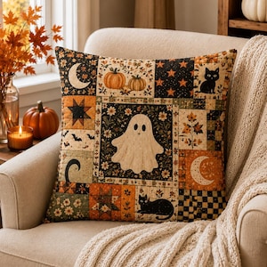 May include: A square throw pillow featuring a patchwork design with various Halloween-themed illustrations including a central white ghost, black cats, orange pumpkins, crescent moons, stars, bats, and floral patterns in shades of orange, black, and cream.