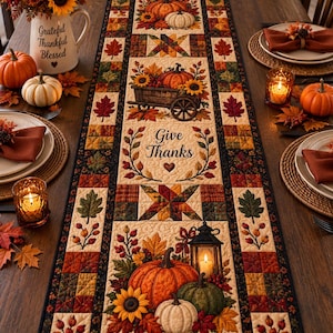 May include: A rectangular fabric table runner featuring a quilted patchwork design with autumn motifs including pumpkins, sunflowers, fall leaves, a wooden cart, and a lantern. The center panel displays the text Give Thanks with a small heart icon.