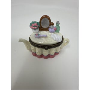 Może przedstawiać: Ozdobne pudełko na bibeloty w kształcie porcelanowego czajniczka. Pokrywka przedstawia miniaturową scenę toaletki z lustrem, flakonami perfum i szczotką do włosów. Pudełko jest kremowe z falistym, różowym wykończeniem.