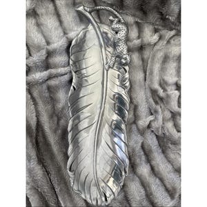Puede incluir: Una bandeja decorativa plateada con forma de pluma grande. Una detallada figura de leopardo está posada en la parte superior de la pluma. La bandeja tiene una superficie texturizada y probablemente es de metal.