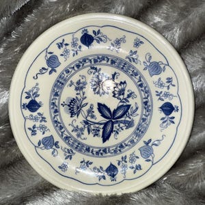 Puede incluir: Un plato redondo de cerámica blanquecina con un diseño floral azul. El diseño presenta flores, hojas y frutas estilizadas. El plato tiene un borde festoneado y un borde detallado, creando una estética clásica y vintage.