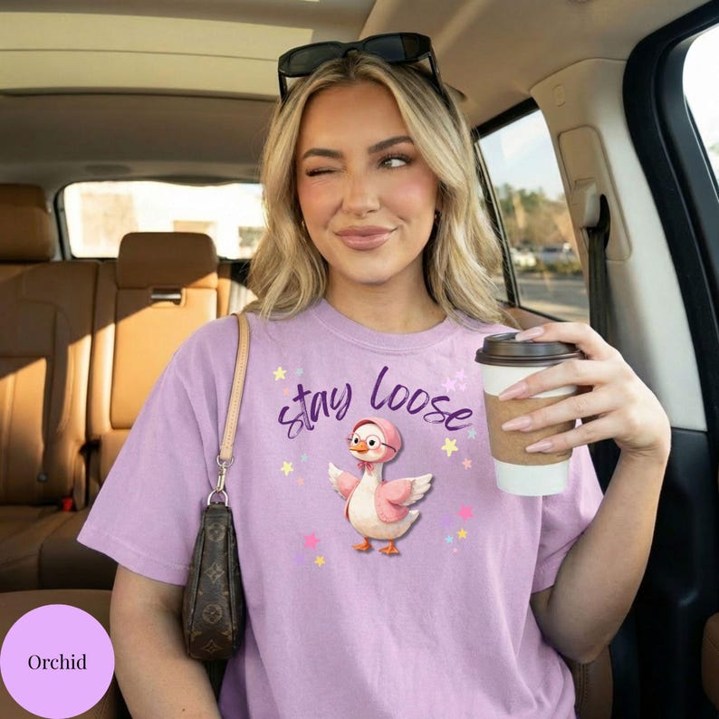 A woman wearing a pink t-shirt with a cartoon duck holding a coffee cup, smiling and looking out the window of a car.
