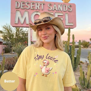 A woman wearing a yellow t-shirt with a cartoon duck and the text "Stay Loose" stands in front of a pink sign for the Desert Sands Motel.
