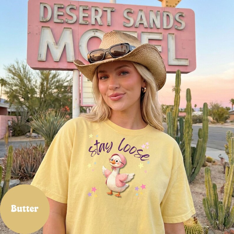 A woman wearing a yellow t-shirt with a cartoon duck and the text "Stay Loose" stands in front of a pink sign for the Desert Sands Motel.