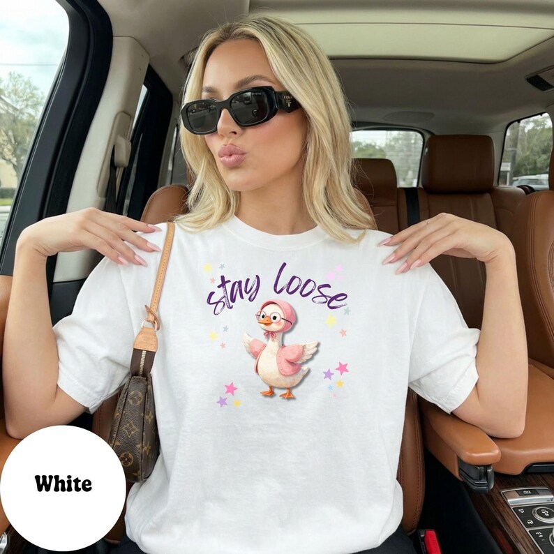 A woman wearing a white t-shirt with a cartoon duck and the text "Stay Loose" is sitting in a car.