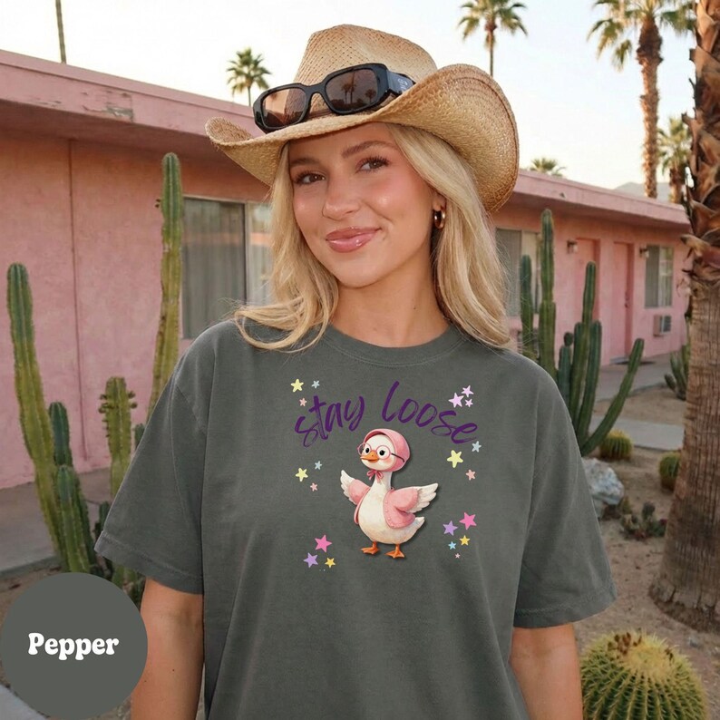 A woman wearing a cowboy hat and sunglasses stands in front of a pink building with palm trees in the background. She is wearing a gray t-shirt with a cartoon duck and stars on it.