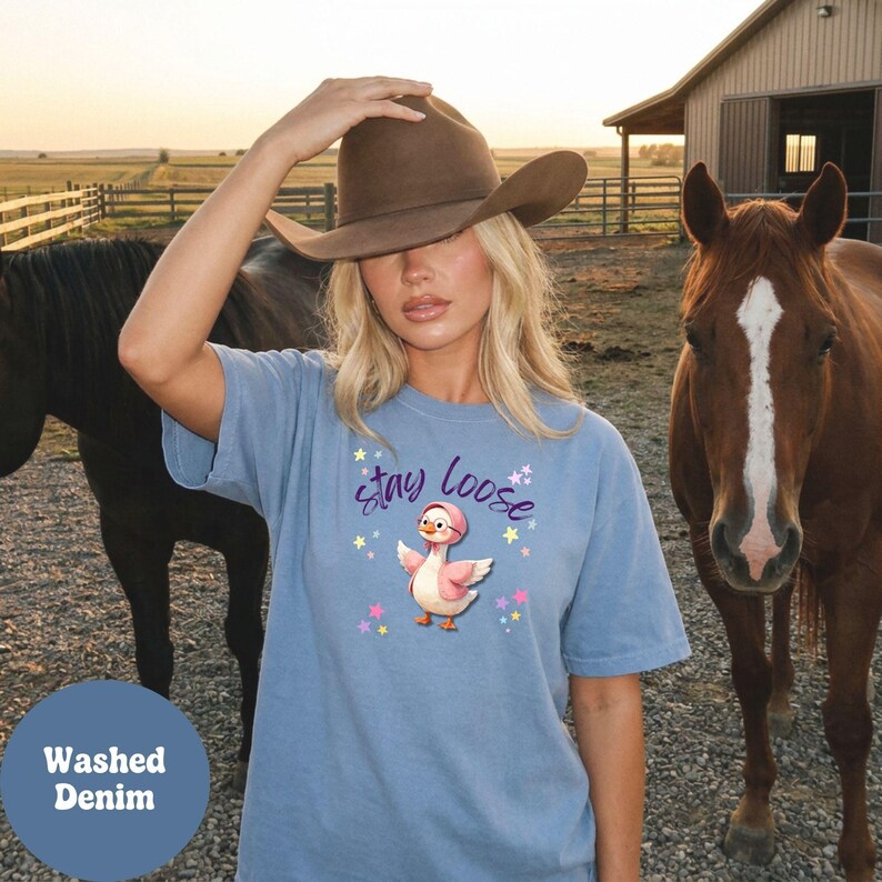 A woman wearing a brown hat and a blue t-shirt with a cartoon duck stands next to two horses in a rural setting.