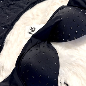 May include: A black two-piece bikini set featuring molded cups and matching bottoms, both adorned with small, evenly spaced black studs. A white tag with the black text NB COUTURE is attached to the center of the bikini top.