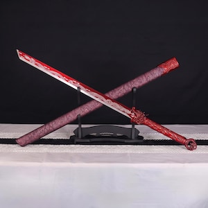 Puede incluir: Una espada roja con un motivo de dragón en la hoja y el mango, expuesta sobre un soporte negro junto a una vaina roja a juego con un intrincado patrón en relieve. La espada y la vaina están dispuestas en forma de cruz sobre el soporte.