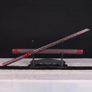 May include: A katana sword with a long, slender blade featuring intricate etched scrollwork patterns. The handle and the matching cylindrical scabbard are finished in a deep, textured maroon red with ornate metallic red accents at the ends.