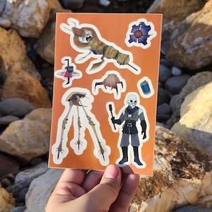 Puede incluir: Una hoja de pegatinas naranja con varias pegatinas de personajes de un videojuego. Las pegatinas incluyen una máquina con forma de trípode, un personaje con atuendo gris y negro y otros diseños relacionados con el juego. Las pegatinas tienen bordes blancos.