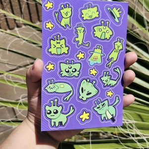 May include: A purple sticker sheet featuring various cartoon alien designs in shades of green and yellow. The aliens have different shapes and expressions, with some holding objects. The sheet is held in a hand.