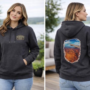 May include: Charcoal gray hoodie with a front pocket and a hood. The front features a patch with the text "WINDOW SEAT CLUB". The back displays a scenic graphic of a landscape. The model is wearing blue jeans.