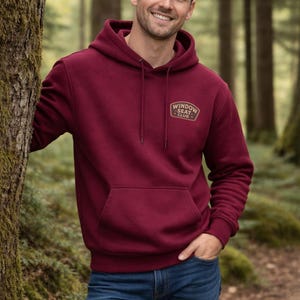 May include: A burgundy hoodie with a front pocket and drawstring hood. The hoodie features a small logo that reads "WINDOW SEAT CLUB" in gold lettering. The person in the image is wearing blue jeans and is standing in a forest setting.