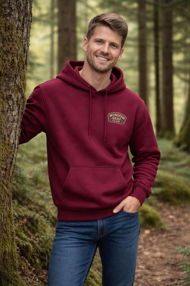 May include: A burgundy hoodie with a front pocket and drawstring hood. The hoodie features a small logo that reads "WINDOW SEAT CLUB" in gold lettering. The person in the image is wearing blue jeans and is standing in a forest setting.