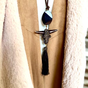 May include: A long necklace featuring a dark brown suede cord, a smooth oval black and white agate stone, and a central pendant shaped like a steer skull with a dark metallic finish and a textured surface, finished with a dark brown tassel.