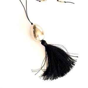 May include: A black tassel necklace with a silver-toned pendant. The pendant is an oval shape with a textured surface. The necklace has a black cord and small silver-toned accents.