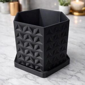 May include: A black, geometric planter with a square base and a textured, triangular pattern. The planter is made of a matte material and sits on a matching square base. The design is modern and minimalist.