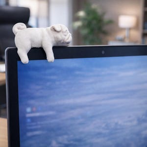 May include: A white pug dog webcam cover resting on top of a laptop. The pug is in a lying down position, with its front paws hanging over the edge of the laptop. The laptop screen displays a blue and white image.