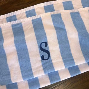 May include: A folded blue and white striped towel with a navy blue letter "S" embroidered on it. The towel has thick, vertical stripes and a white border. The towel is made of a soft, absorbent material.
