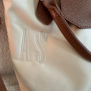 以下が含まれることがあります： 茶色の革のアクセントが付いた、クリーム色のトートバッグ。バッグは、トップハンドル、フラップクロージャー、白で刺繍されたイニシャル「AS」が特徴です。バッグの金具はゴールドトーンです。