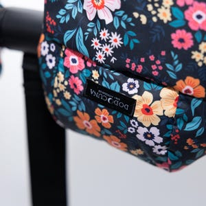 May include: Close-up of a floral print accessory. The accessory features a dark background with colorful flowers in shades of pink, orange, yellow, and white, with teal leaves. The label reads "DODO CUNA".
