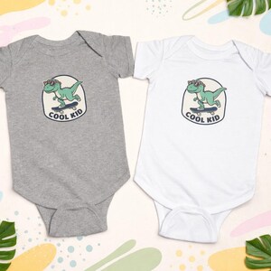 May include: Two baby onesies, one gray and one white, each featuring a cartoon dinosaur wearing sunglasses and skateboarding. The text "COOL KID" is below the dinosaur. The background includes tropical leaves.