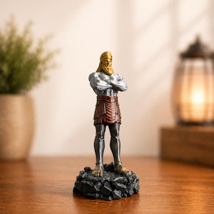 May include: A small figurine of a muscular man with a golden beard and hair, wearing a brown skirt and silver armor. The figure stands on a black, rocky base. The statue has its arms crossed and is a decorative item.