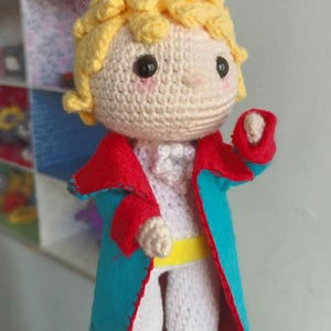 Puede incluir: Muñeca de ganchillo hecha a mano de un personaje con cabello amarillo, que lleva una camisa blanca, pantalones grises y un abrigo azul con ribetes rojos. La muñeca tiene ojos negros de botón y un cinturón amarillo. La muñeca mide aproximadamente 20 cm de altura.