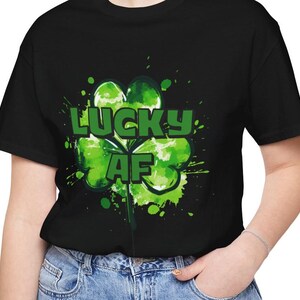 Puede incluir: Camiseta negra con un diseño de trébol de cuatro hojas verde y las palabras "LUCKY AF" en verde. El trébol tiene un efecto acuarela con salpicaduras de pintura verde. La camiseta la lleva una persona con vaqueros azules.