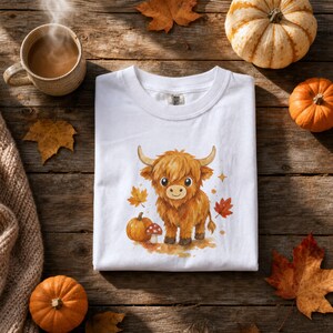 May include: A white t-shirt featuring a watercolor-style illustration of a fluffy brown Highland cow standing next to a small orange pumpkin and a red-and-white spotted mushroom, surrounded by scattered autumn leaves in shades of gold and red.