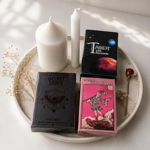 May include: Three tarot card decks on a round tray: a black box labeled Occult Tarot, a black box labeled Tarot with Keywords featuring space imagery, and a pink box labeled Pink Tarot for Beginners. Two white candles and dried flowers are included.