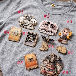 May include: A collection of enamel pins featuring fantasy themes, including dragons, books, and a cat with bat wings. The pins are in various shapes and colors, with text that reads "Fourth Wing", "Onyx Storm", "Iron Flame", and "Books Are Magic".