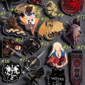 May include: A collection of acrylic pins featuring characters and symbols from a fantasy series. Designs include dragons, a raven, a direwolf, and portraits of characters. The pins are in various colors, including red, black, and gold.