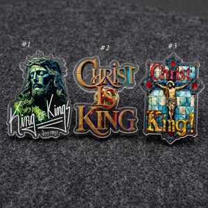 May include: Three religious-themed pins with clear borders. The first pin features a portrait of Jesus with the text "King of Kings". The second pin says "Christ is King" in gold and blue. The third pin depicts Jesus on a cross with the text "Christ King!"