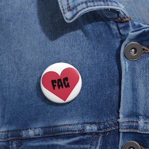 May include: A white button with a red heart design and the word "FAG" in black lettering. The button is pinned to a blue denim jacket.