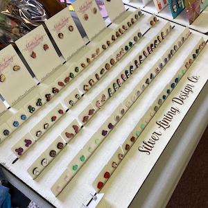 Puede incluir: Un tablero de madera blanco con filas de pendientes de madera de colores. Los pendientes están dispuestos en pares y se muestran en pequeñas tarjetas blancas. El tablero tiene el texto "Silver Lining Design Co." impreso en él.