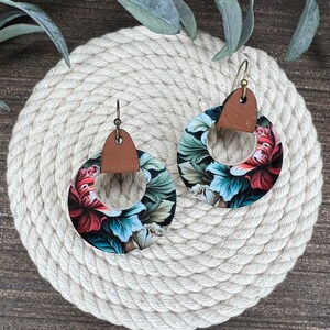 May include: A pair of floral print earrings with brown leather accents. The earrings are round and have a large floral design with shades of red, green, and blue.