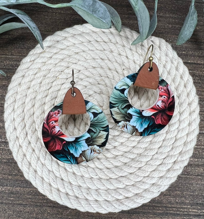May include: A pair of floral print earrings with brown leather accents. The earrings are round and have a large floral design with shades of red, green, and blue.
