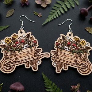 Può includere: Orecchini in legno con un design a carriola riempita di funghi, fiori e felci. Gli orecchini hanno un gancio d'argento e sono decorati con un teschio e dettagli di fantasmi. Gli orecchini sono di colore marrone chiaro.