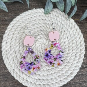 May include: A pair of floral print earrings with pink accents. The earrings are made of polymer clay and feature a floral pattern with purple and pink flowers. The earrings are suspended from silver hooks.