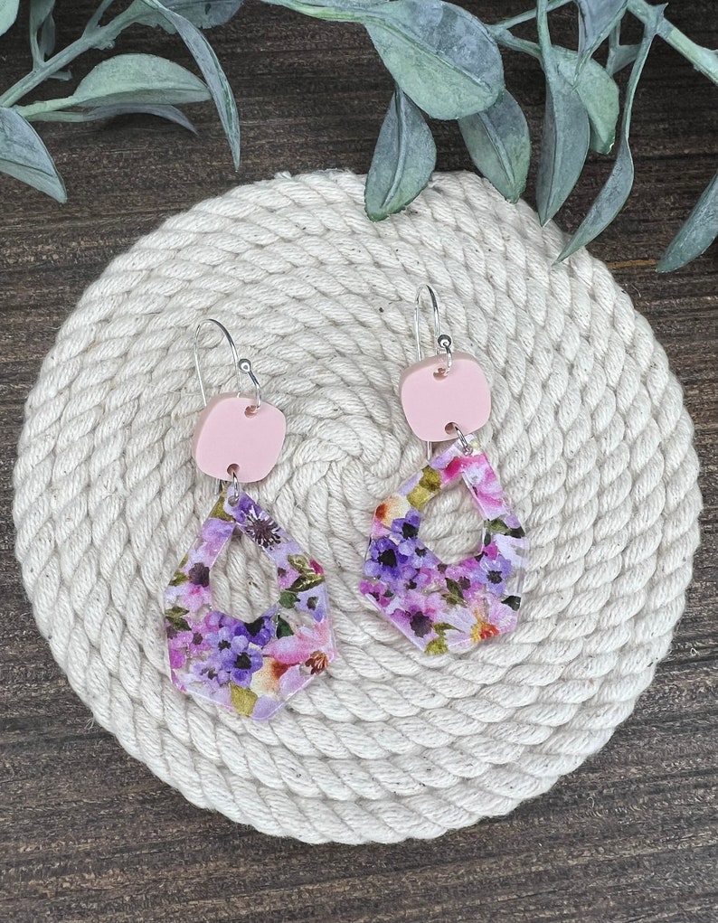 May include: A pair of floral print earrings with pink accents. The earrings are made of polymer clay and feature a floral pattern with purple and pink flowers. The earrings are suspended from silver hooks.