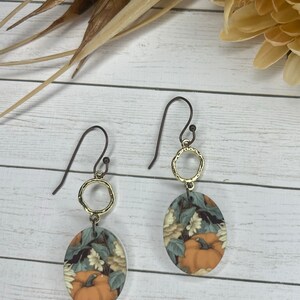May include: A pair of dangle earrings with a gold oval charm featuring a vintage-style illustration of pumpkins and flowers. The charms are attached to a gold ring and a brown wire hook.