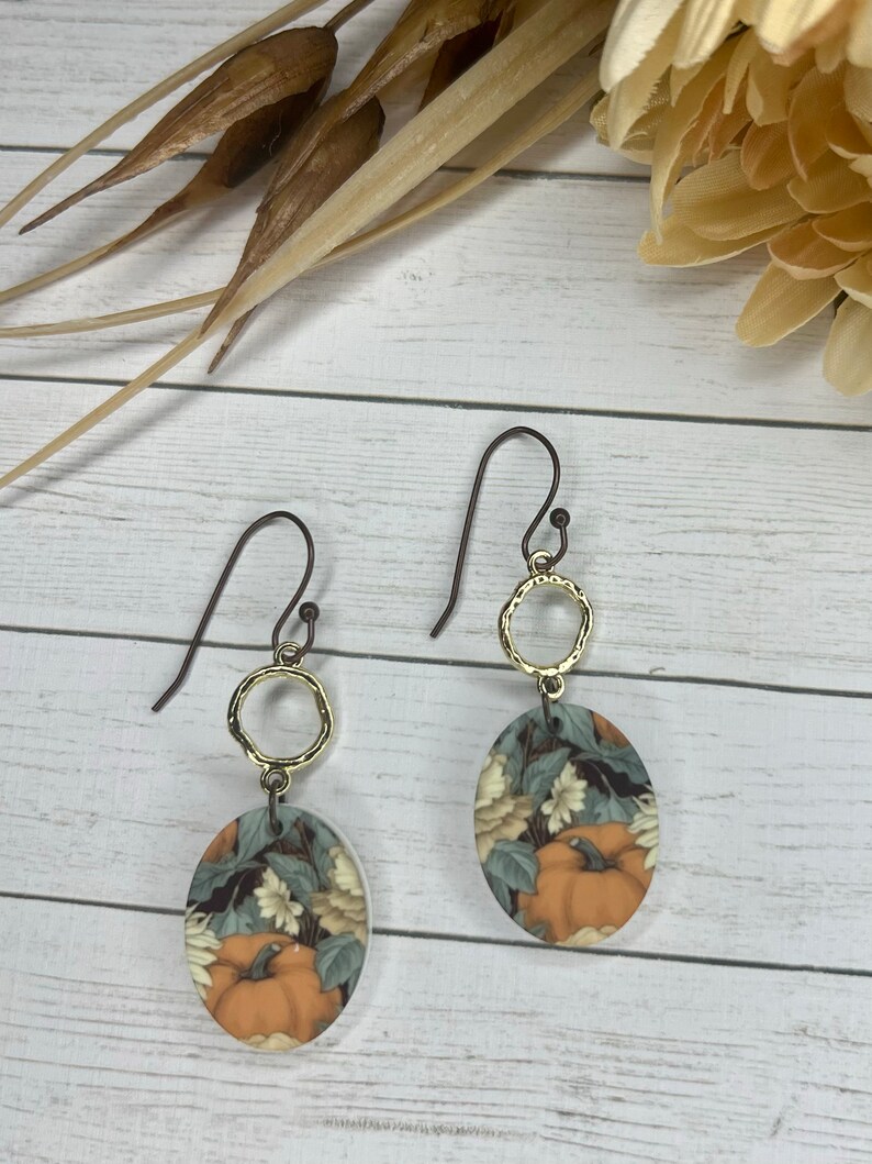 May include: A pair of dangle earrings with a gold oval charm featuring a vintage-style illustration of pumpkins and flowers. The charms are attached to a gold ring and a brown wire hook.