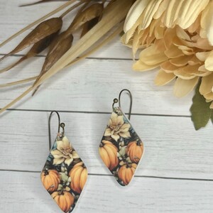 May include: A pair of diamond-shaped earrings with a fall-themed design featuring pumpkins, flowers, and leaves. The earrings are made of metal and have a silver finish.