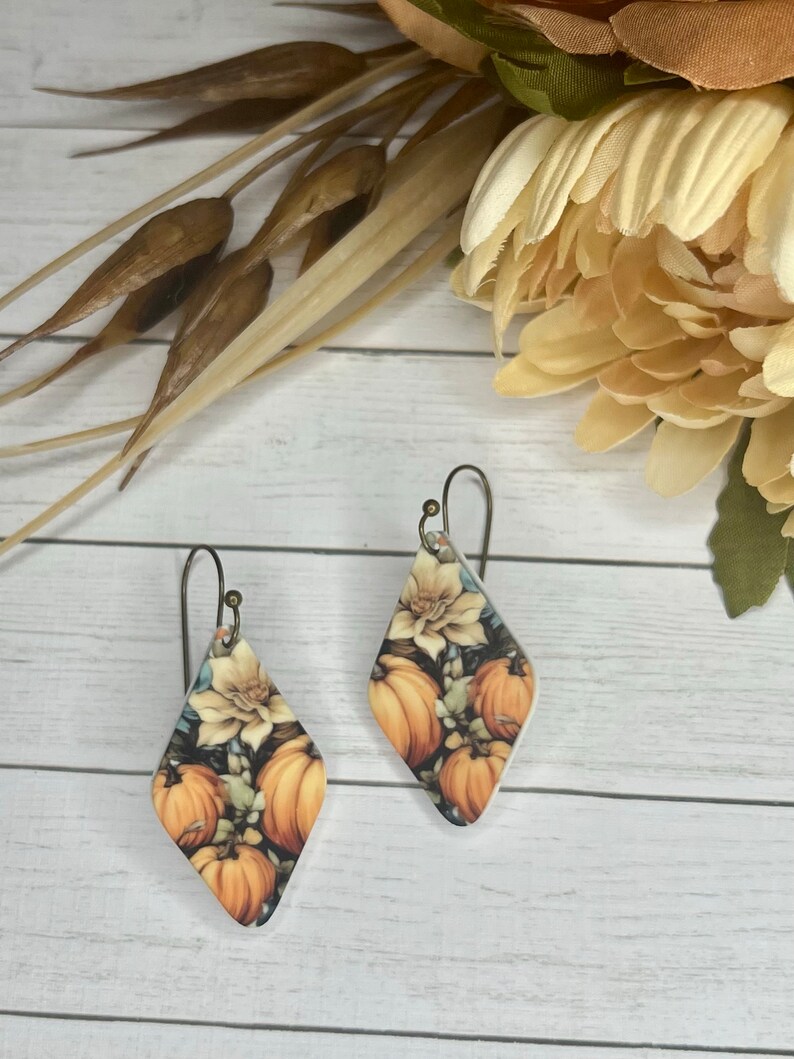 May include: A pair of diamond-shaped earrings with a fall-themed design featuring pumpkins, flowers, and leaves. The earrings are made of metal and have a silver finish.