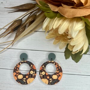 May include: A pair of round, dangle earrings with a black background and a pattern of orange pumpkins, leaves, and flowers. The earrings have a gray top and a silver ring in the center.